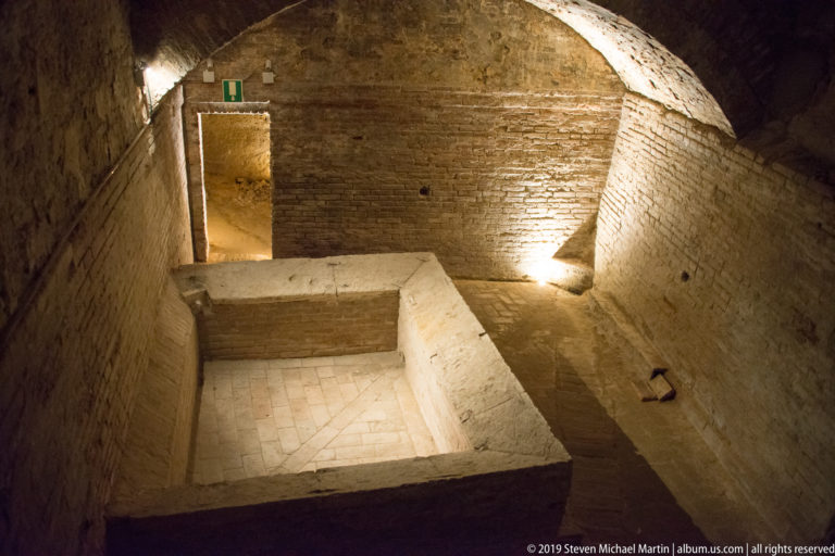 Brick-vaulted underground chamber with a large rectangular masonry basin and tiled floor lit by spotlights.