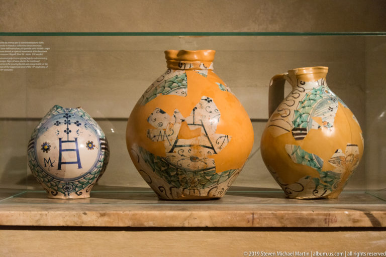 Three glazed ceramic vessels on a shelf, including a blue-and-white round jar and two ochre figural jars.