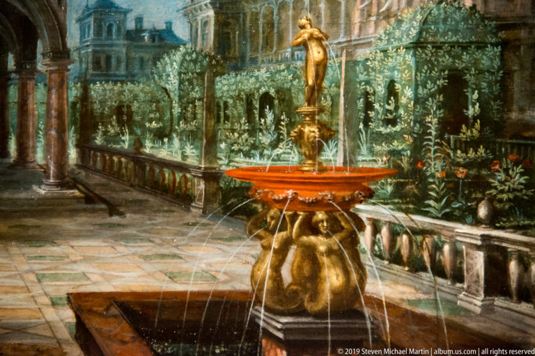 Gilded sculptural fountain spraying arcs of water in a formal garden bordered by stone balustrades and architecture.