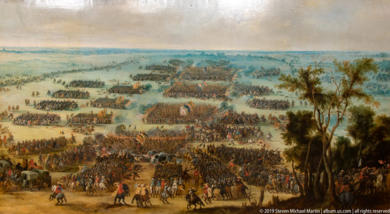 Wide painted battlefield crowded with infantry and cavalry formations stretching across a hazy plain.