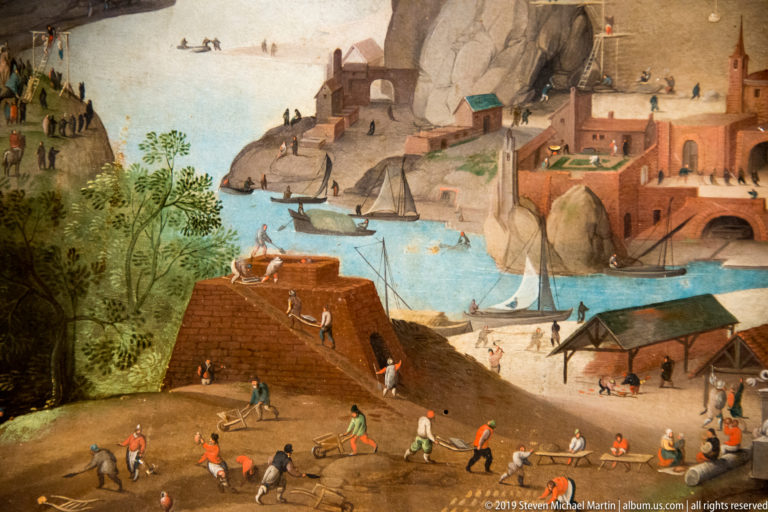 Laborers carry loads along a road beside blue water, boats, brick buildings, and rocky slopes.