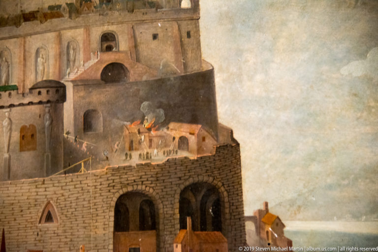 Small building burns beside massive stone fortifications as smoke rises and tiny figures gather nearby.