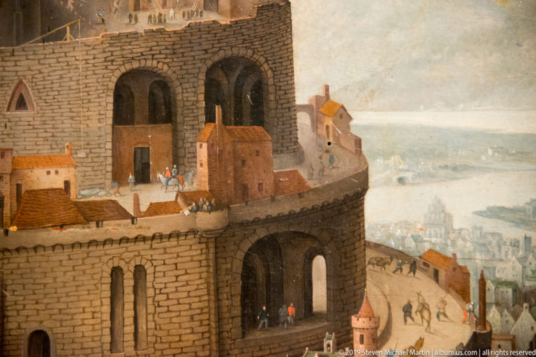 Large painted masonry tower with arched openings, tiny figures, steep road, distant city, and water.