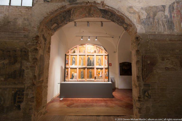 Rough stone arch with faded wall paintings framing a brightly lit multi-panel artwork on a museum plinth beyond.