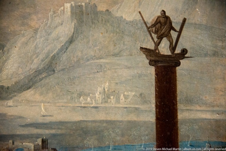 Tall brown column topped by a standing figure, with pale mountains, water, boats, and distant buildings.