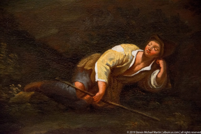 Reclining man asleep on dark ground, head resting on one hand and a long staff across his body.