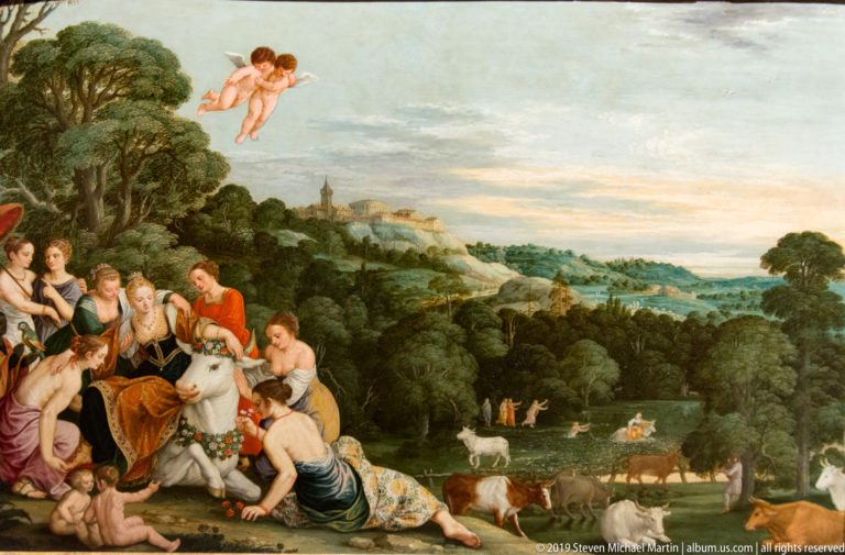 Wide painted landscape with women around a garlanded white bull, grazing animals, woodland, and a distant town.