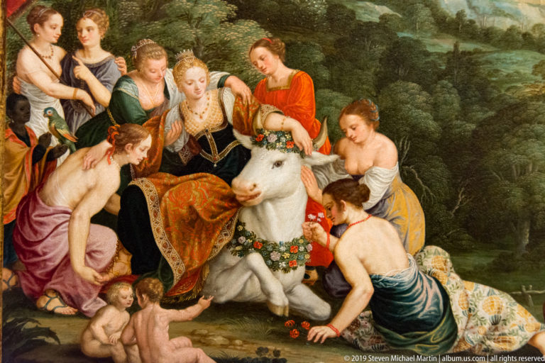 Women and nude children gather around a flower-garlanded white bull in a wooded landscape.