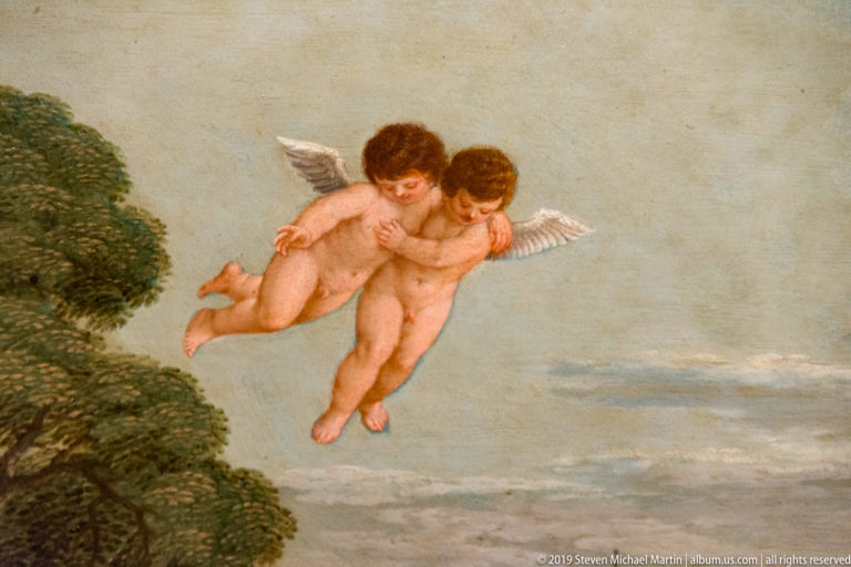 Two nude winged putti embracing in flight above a leafy tree against a pale sky.