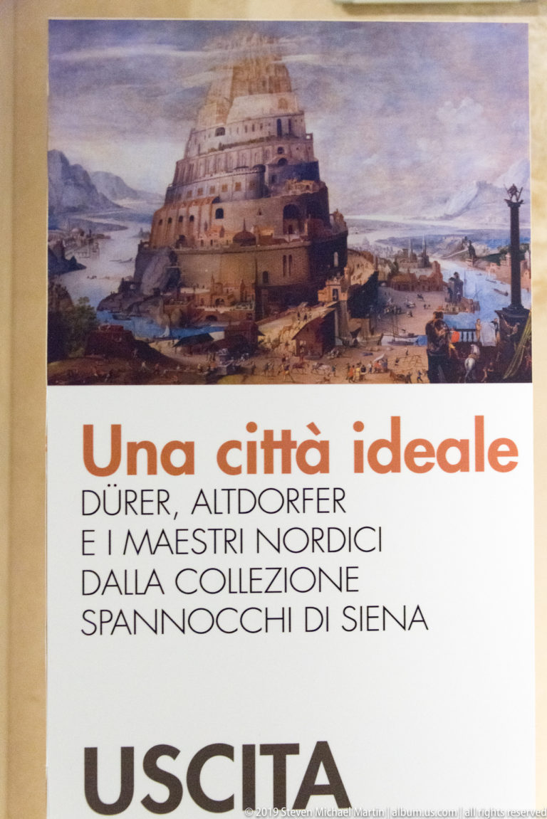 Museum exit panel with a painted terraced city above Italian exhibition text and the large word “USCITA.”