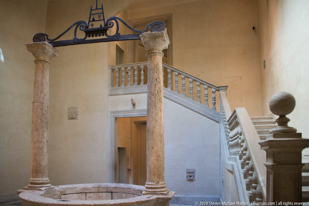 Stone courtyard with two columns, circular basin, balustraded stair, doorway, and pale plaster walls.