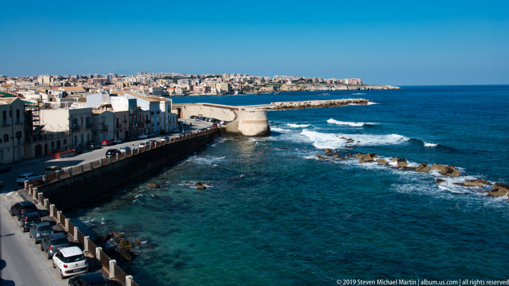 Elevated view along Ortigia’s seafront with pale buildings, coastal road, seawall, rocky shallows, surf, and blue sea.