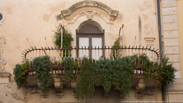 Iron balcony densely filled with trailing plants beneath an arched window on a weathered ocher façade.