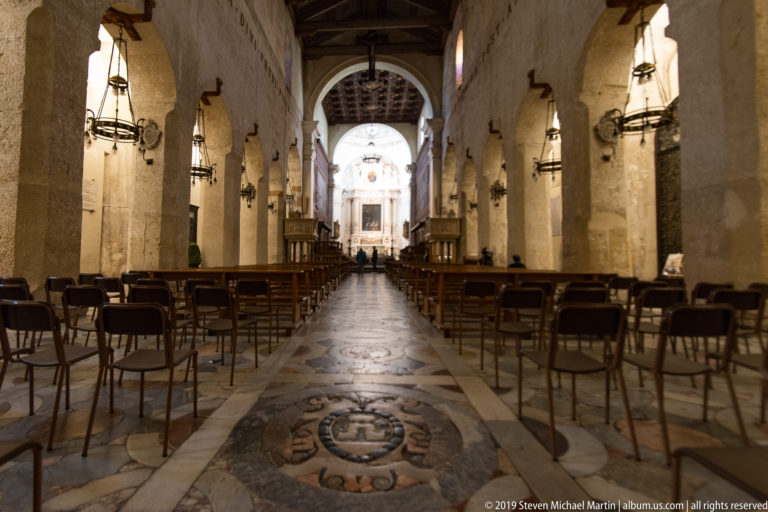 Central aisle of Syracuse Cathedral with heraldic marble floor inlay, rows of chairs, stone supports, and distant altar.