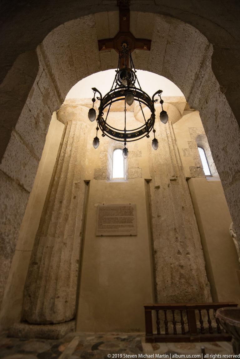 Low-angle view inside Syracuse Cathedral of a massive pale stone pier and arch with chandelier, narrow windows, and wall plaque.