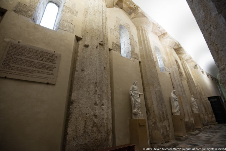 Tall pale masonry inside Syracuse Cathedral with a commemorative plaque, narrow high windows, and statues on pedestals.