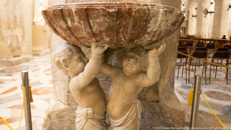 Two carved putti support a large reddish veined holy-water basin against pale masonry inside the church.