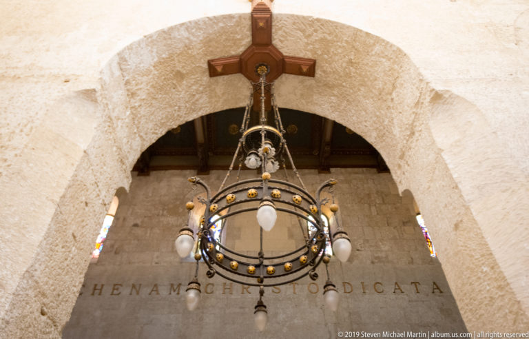Large circular chandelier hangs beneath a rough stone arch and wooden cross, with small stained-glass windows nearby.