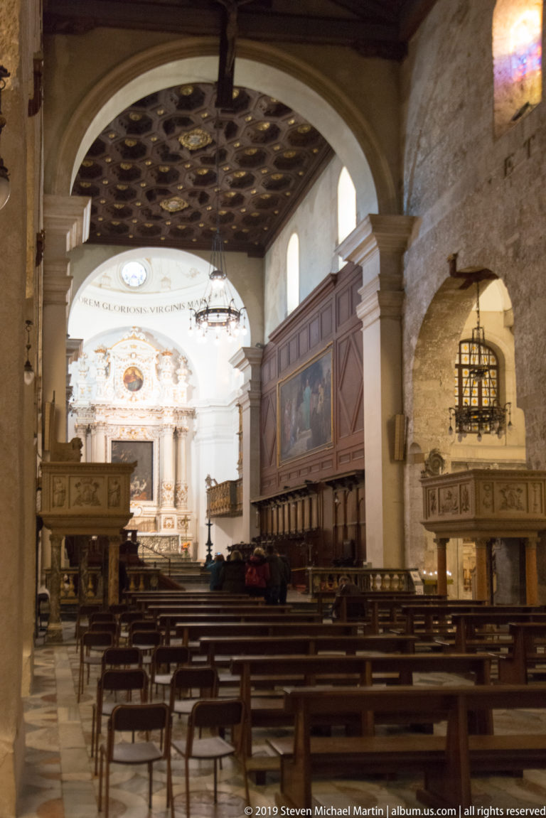 Wide cathedral interior with rows of chairs, stone arches, dark choir enclosures, coffered ceiling, and ornate altar.
