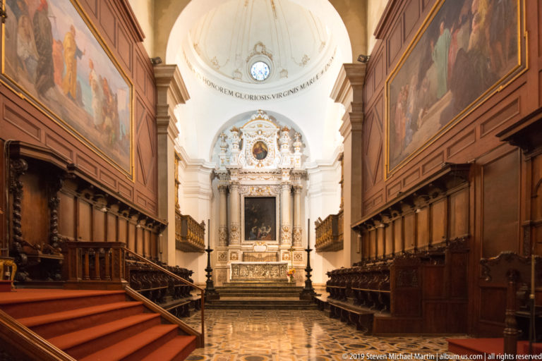 Symmetrical dark wooden choir stalls and painted panels lead toward an ornate altar in a bright apse.