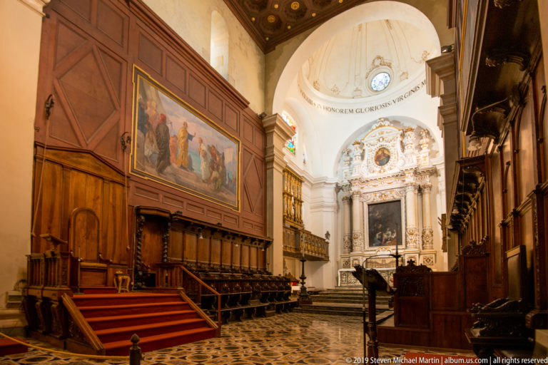 Dark wooden choir stalls flank a central aisle leading to an ornate pale altar in a vaulted sanctuary.