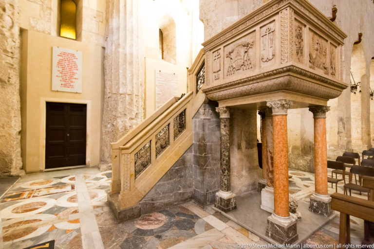 Raised carved stone pulpit with staircase, relief panels, and colored supporting columns inside the cathedral.