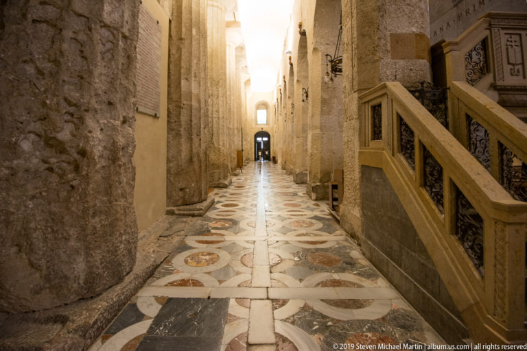 Narrow cathedral side aisle with massive stone supports, patterned marble floor, and a carved stair at right.