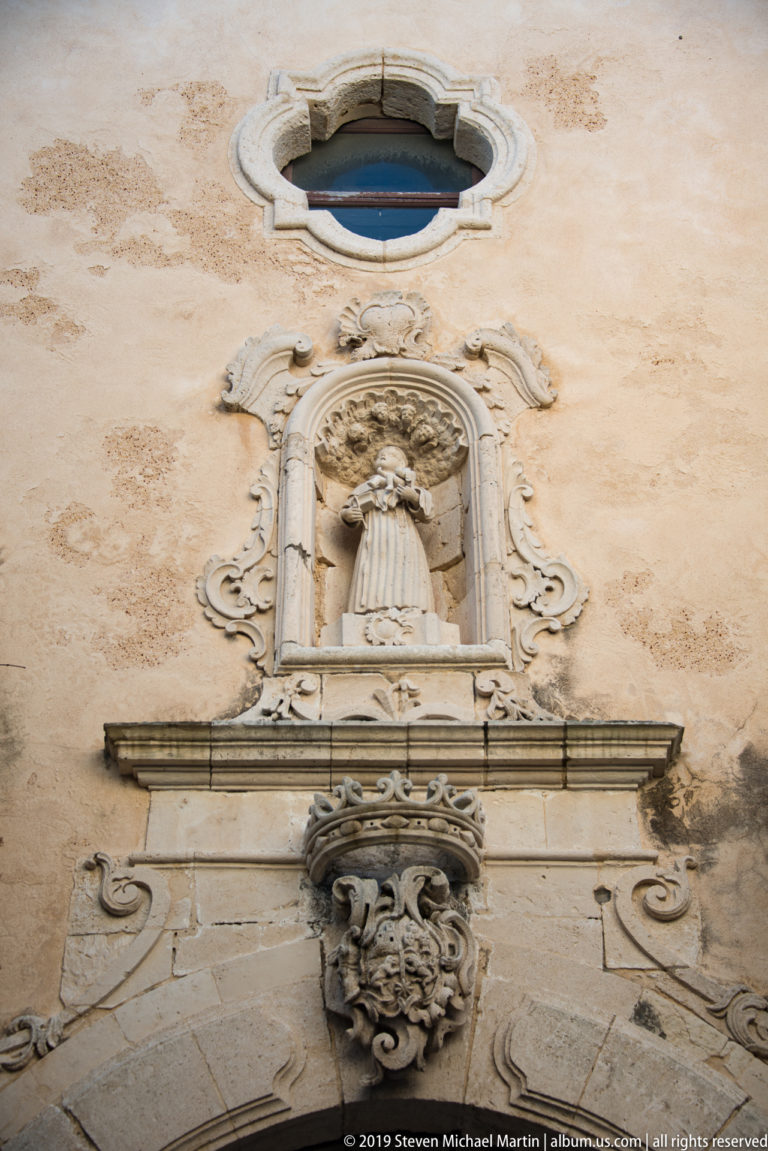 Ornate stone devotional niche beneath an oval window, with scrolling decoration and a large grotesque mask corbel below.
