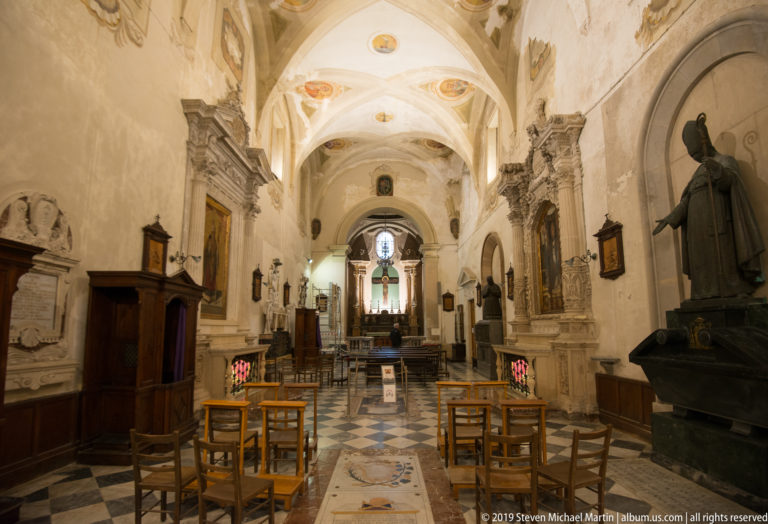Long central view inside Syracuse Cathedral with pale stone walls, monuments, chairs, hanging lamps, and a distant altar.