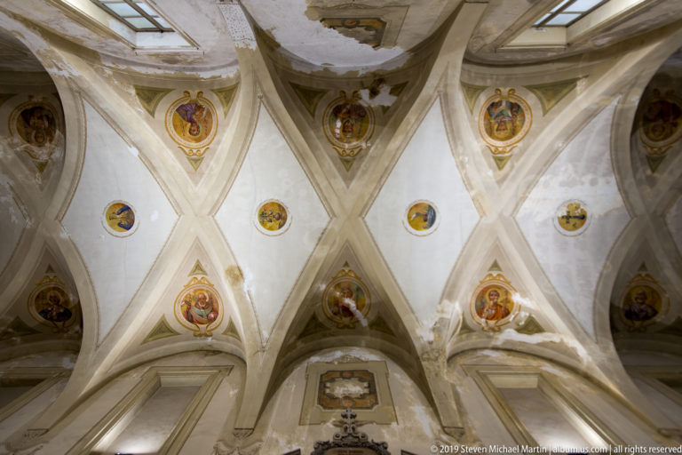 Upward view of intersecting pale vault ribs with painted religious medallions and patches of worn plaster.