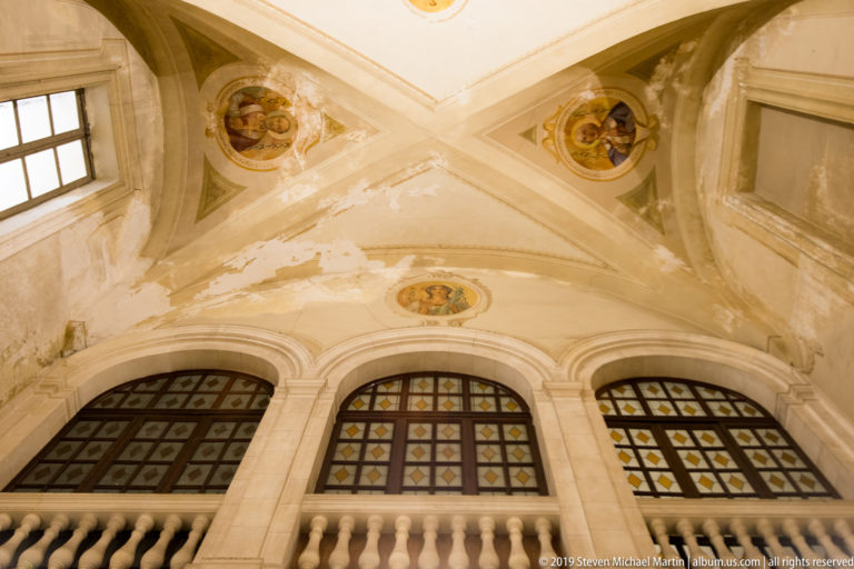 Pale groin vault with small painted medallions above three patterned windows and a balustrade, with areas of peeling plaster.