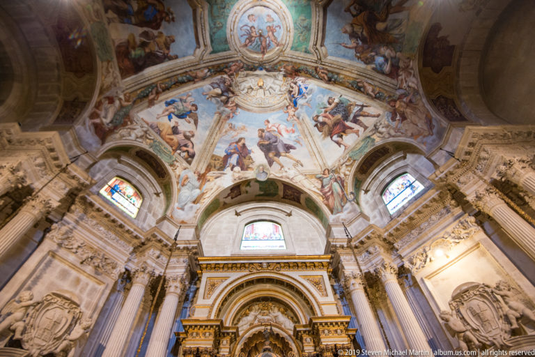 Steep upward view of narrative ceiling frescoes, ornate moldings, windows, and the upper portion of a church altar.