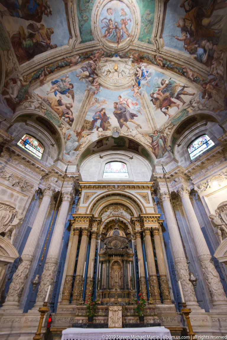 Ornate church altar with paired columns beneath a brightly frescoed vaulted ceiling and stained-glass windows.
