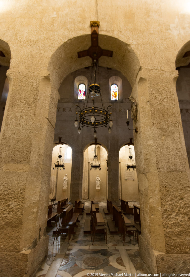 Massive ancient stone piers and broad arches inside Syracuse Cathedral, with chandeliers, chairs, stained glass, and side altars.