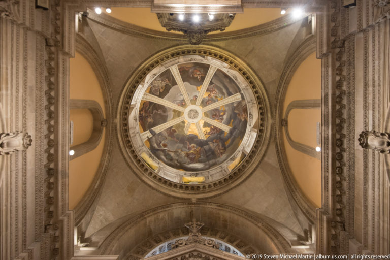 Frescoed circular dome centered above broad arches and ornate architectural moldings in a church interior.