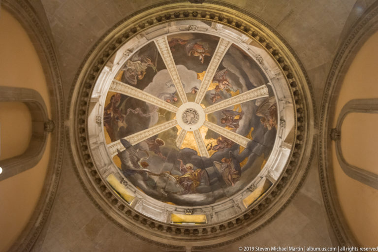 Upward view of a circular frescoed dome divided by pale radial ribs into painted figurative scenes.