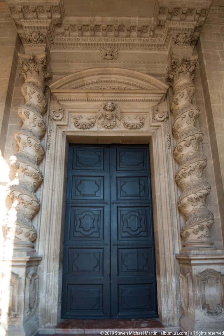 Dark paneled double doors framed by an ornate pale stone Baroque portal with twisted columns and carved ornament.