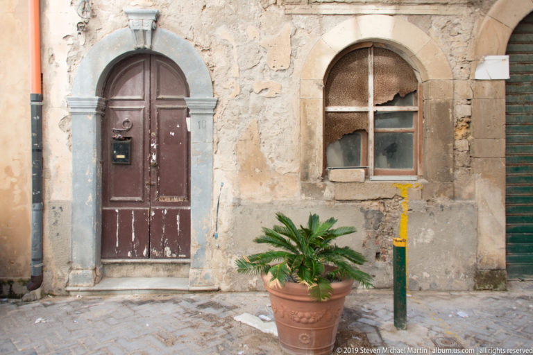 Worn arched wooden door beside a small window in patched stonework, with a potted cycad and utility post.