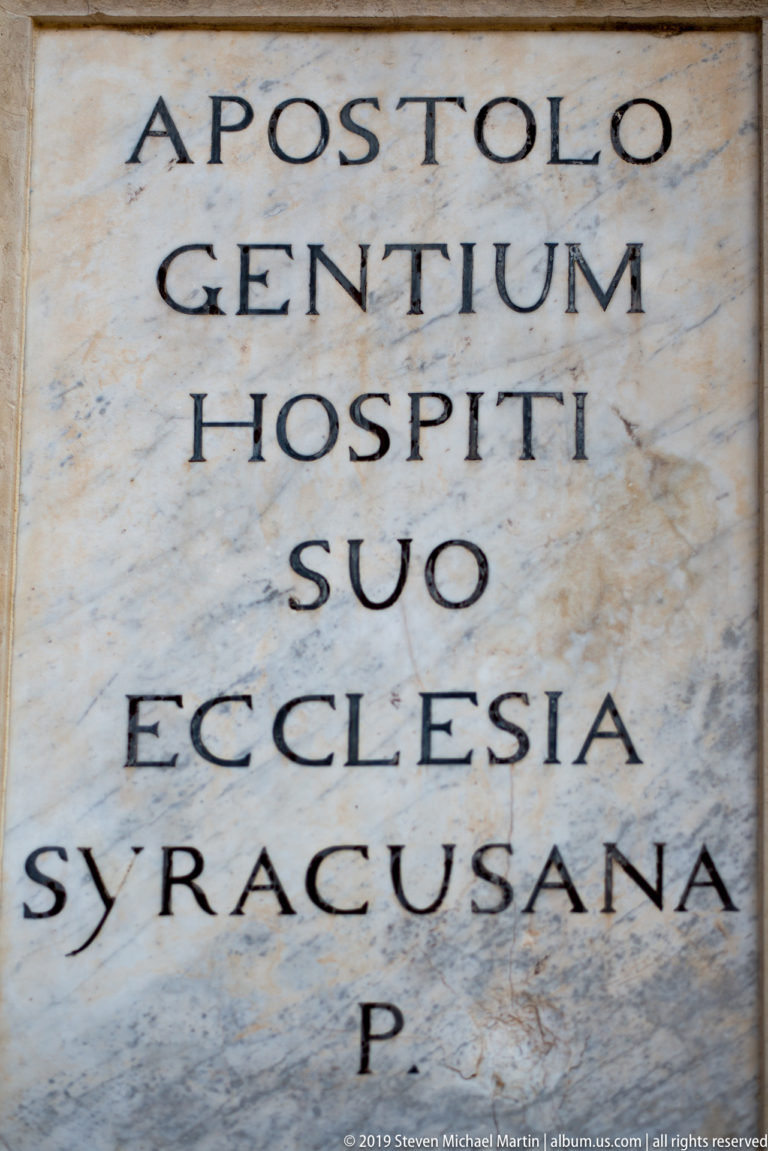 Close-up of a pale marble slab with black Latin capitals reading “APOSTOLO GENTIUM HOSPITI SUO ECCLESIA SYRACUSANA.”