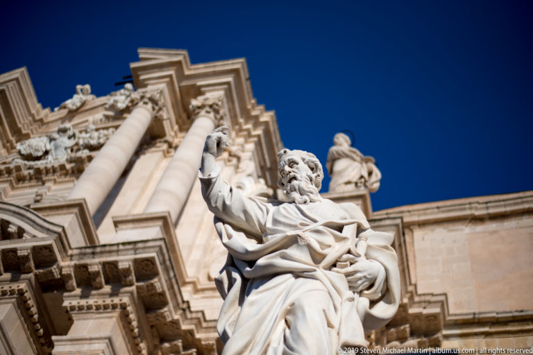 Low-angle view of the monumental Saint Paul statue before the columns and sculptural Baroque façade of Syracuse Cathedral.