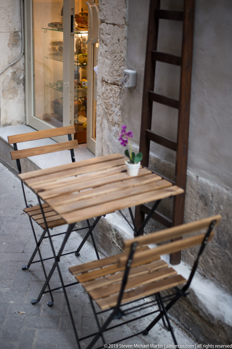 Folding wooden café tables and benches beside a rough stone wall and open doorway, with a small potted flower.