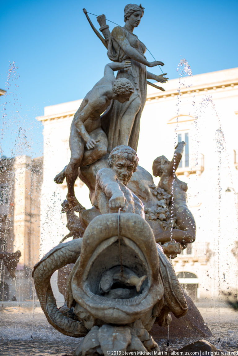 Stone figures and a coiling sea creature in the Fountain of Diana, with water jets against a bright blue sky.