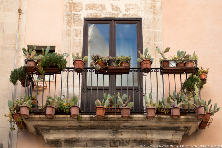 Small wrought-iron balcony beneath a wooden window, densely lined with terracotta pots of succulents and trailing plants.