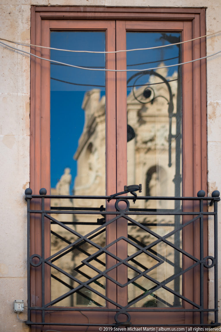 Ornate pale stone architecture reflected in a blue window above a black wrought-iron balcony railing and utility wires.