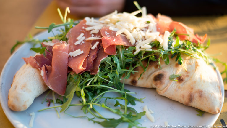 Folded blistered flatbread filled with arugula, thin cured ham, and shaved pale cheese on a plate.