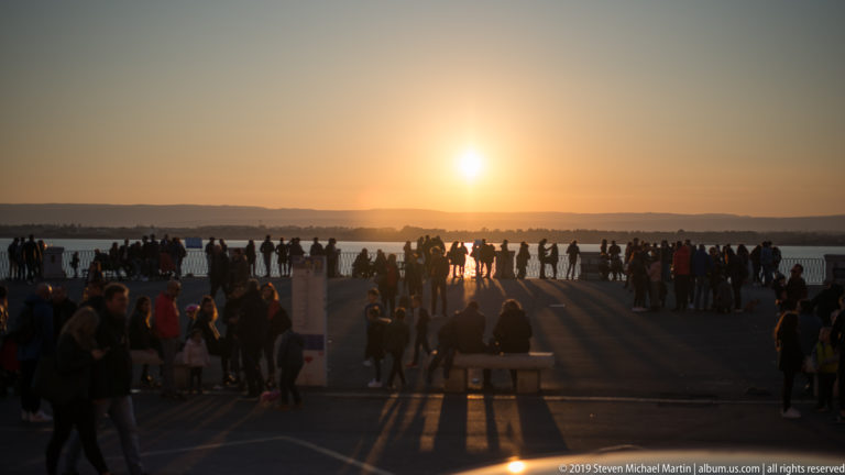Visitors crowd a broad waterfront promenade at sunset, casting long shadows toward the camera beneath a golden sky.