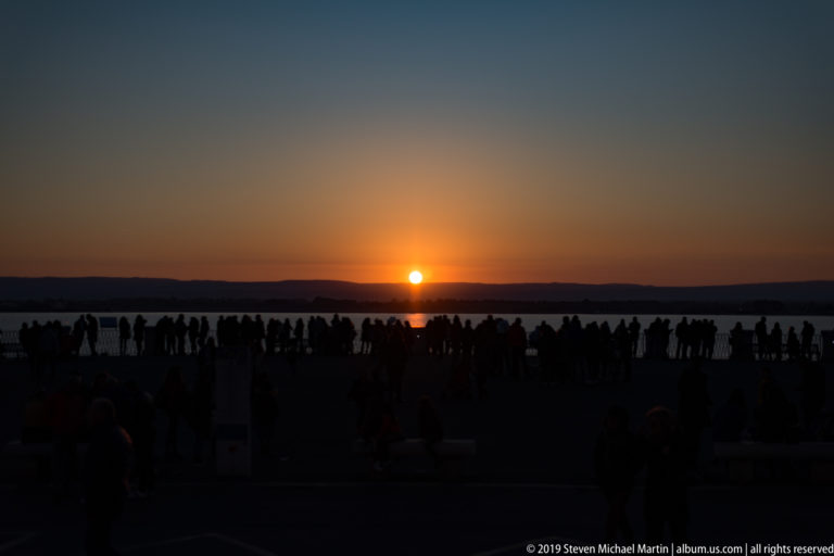 Silhouetted crowd gathered at a waterfront overlook as the sun sets on the horizon beyond Ortigia.