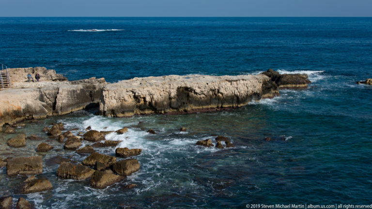 Low limestone rock ledge extending into deep blue sea with people on the rocks and white waves breaking around it.