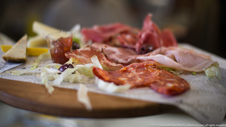 Close-up of sliced cured meats, sausage, pale cheese, and accompaniments arranged on a round wooden serving board.