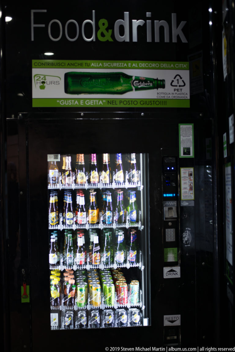 Illuminated food-and-drink vending machine stocked with bottles and packaged items in a dark storefront.